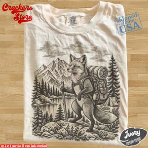 Può includere: Una t-shirt color avorio con un'illustrazione grafica marrone scuro di una volpe che fa escursionismo in un paesaggio montano. La volpe indossa uno zaino, porta un bastone da passeggio ed è vicino a un lago, pini e una tenda.