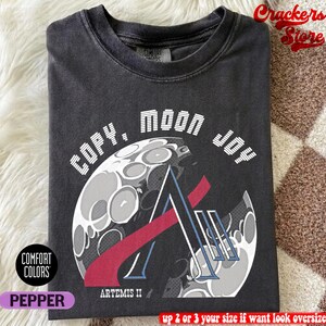 以下が含まれることがあります： 「COPY. MOON JOY」の文字と月のグラフィック、Artemis IIのロゴが入った濃いグレーのTシャツ。折りたたまれたシャツにはComfort Colorsのタグが付いています。色はペッパーです。