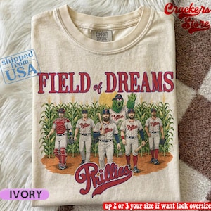 Op de afbeelding: Een ivoorkleurig t-shirt met ronde hals en een afbeelding van vijf honkbalspelers en een groene mascotte in een maïsveld. De tekst FIELD of DREAMS en Phillies staat in rode letters op het shirt. Het shirt heeft een Comfort Colors label.