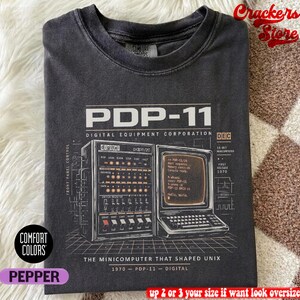 May include: A dark gray t-shirt with a graphic of a PDP-11 minicomputer. The text on the shirt includes "PDP-11 DIGITAL EQUIPMENT CORPORATION" and "THE MINICOMPUTER THAT SHAPED UNIX". The shirt is labeled "Comfort Colors" and "Pepper".