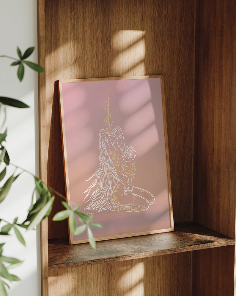 May include: A framed print with a pink background and a white line drawing of a person with long hair, kneeling with their arms raised above their head. The person is adorned with floral details and a rose.