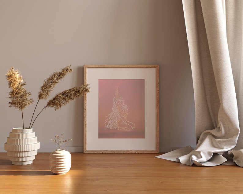 May include: A framed print with a minimalist line drawing of two figures embracing, set against a pink and orange gradient background. The print is displayed on a wooden floor with a vase of dried pampas grass and a small vase of flowers in the foreground.