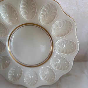 May include: Off-white ceramic oyster serving plate with a scalloped edge and gold trim. The plate features 12 oval indentations with a leaf-like design, surrounding a central circular area.