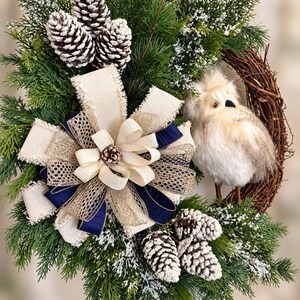 May include: A decorative wreath featuring green pine branches with white-tipped needles, clusters of frosted pinecones, and a large multi-layered bow made of cream-colored burlap, gold mesh, and navy blue ribbon, accented by a small plush owl.