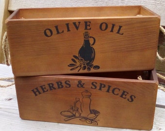 Puede incluir: Dos cajas de almacenamiento de madera con asas de cuerda. La caja superior tiene el texto "Olive Oil" y una silueta negra de una botella de aceite de oliva y aceitunas. La caja inferior tiene el texto "Herbs & Spices" y una silueta negra de un tarro de especias, una cuchara y hierbas.