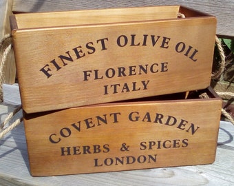 May include: Two wooden boxes with rope handles, stacked on top of each other. The top box reads "FINEST OLIVE OIL FLORENCE ITALY" in black lettering. The bottom box reads "COVENT GARDEN HERBS & SPICES LONDON".