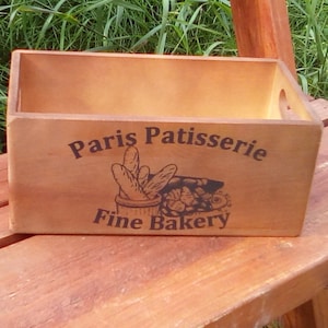 Puede incluir: Una caja rectangular de madera clara con la inscripción "Paris Patisserie Fine Bakery". La caja tiene un diseño sencillo y asas recortadas en los laterales, ideal para almacenamiento.