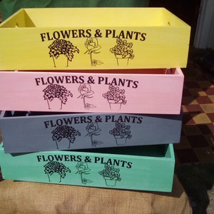 May include: Four wooden boxes stacked on top of each other. The boxes are painted in different colours: yellow, pink, grey, and green. Each box has the words "FLOWERS & PLANTS" printed on the front, along with a black silhouette of a flower and a plant in a pot.