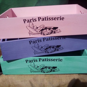 Puede incluir: Tres cajas de almacenamiento de madera en colores pastel: rosa, morado y turquesa. Cada caja rectangular tiene asas recortadas y el texto "Paris Patisserie" con una ilustración de pan y pastelería. Las cajas están apiladas.