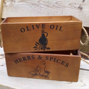 May include: Two wooden rectangular boxes stacked. The top box is labelled "OLIVE OIL" with a black illustration of a bottle and olives. The bottom box reads "HERBS & SPICES" with a black illustration of a mortar and pestle. Each box has rope handles.