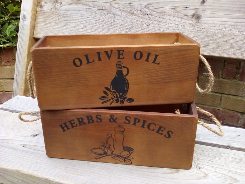 May include: Two wooden rectangular boxes stacked. The top box is labelled "OLIVE OIL" with a black illustration of a bottle and olives. The bottom box reads "HERBS & SPICES" with a black illustration of a mortar and pestle. Each box has rope handles.