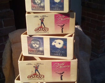 May include: A stack of six wooden boxes with different musical show themes. The boxes are decorated with images of the shows, including Les Miserables, Grease, Dirty Dancing, and The Phantom of the Opera. The boxes are arranged in a pyramid shape.