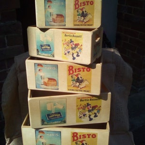 Può includere: Pila di cinque scatole di legno con etichette vintage che mostrano il testo "HOVIS" e "BISTO" insieme a illustrazioni di bambini e pane. Le scatole sono impilate su una superficie di juta.