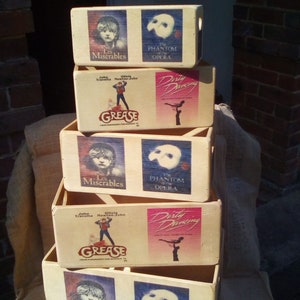 May include: A stack of five wooden boxes with different musical show posters on each side. The posters feature the musicals Les Miserables, Grease, Dirty Dancing, and The Phantom of the Opera.