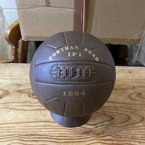 May include: A brown leather soccer ball with gold lettering. The ball says "Portman Road IP1 1884".