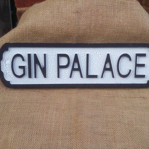 以下が含まれることがあります： 白と黒の看板に、白い背景に黒い文字で「GIN PALACE」と書かれています。看板は、使い古された、アンティークな外観をしています。