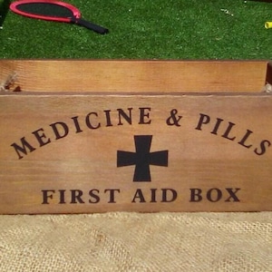 May include: A rectangular wooden box with a medium brown finish and rope handles on the sides. The front features black text that reads MEDICINE & PILLS with a black cross symbol in the centre and the text FIRST AID BOX printed below it.