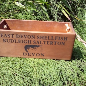 May include: A wooden box with rope handles, featuring the text "East Devon Shellfish Budleigh Salterton Devon" and a black silhouette of a shrimp.