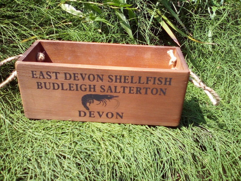 May include: A wooden box with rope handles, featuring the text "East Devon Shellfish Budleigh Salterton Devon" and a black silhouette of a shrimp.