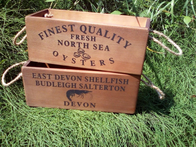 May include: Two wooden boxes with rope handles. The top box has the text "FINEST QUALITY FRESH NORTH SEA OYSTERS" printed on it. The bottom box has the text "EAST DEVON SHELLFISH BUDLEIGH SALTERTON DEVON" printed on it.
