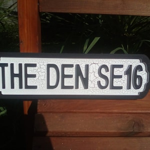 Peut inclure: Un panneau de rue noir et blanc avec le texte "THE DEN SE 16" en lettres blanches sur fond noir. Le panneau a un aspect de peinture craquelée et vieillie.