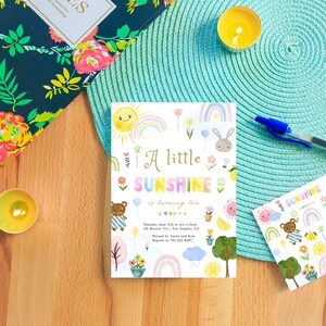 May include: A colorful invitation for a second birthday party with a sun, rainbow, and teddy bear theme. The invitation says "A little sunshine is turning two".