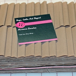 May include: A stack of brown fabric strips, folded and ready for sewing. The fabric is a light brown color and has a smooth texture. A business card with the text "Bags, Quilts And Beyond" and "Marianne Donahue" is placed on top of the fabric.