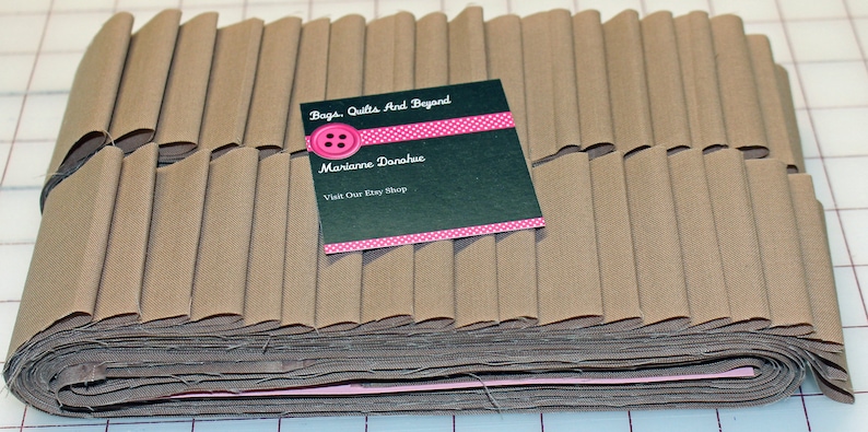May include: A stack of brown fabric strips, folded and ready for sewing. The fabric is a light brown color and has a smooth texture. A business card with the text "Bags, Quilts And Beyond" and "Marianne Donahue" is placed on top of the fabric.