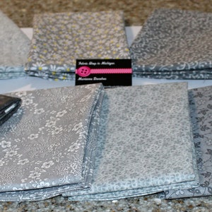 May include: A collection of folded fabric squares in various shades of gray, featuring floral, swirl, and abstract patterns. Some squares have white or yellow accents. A business card is visible in the center.