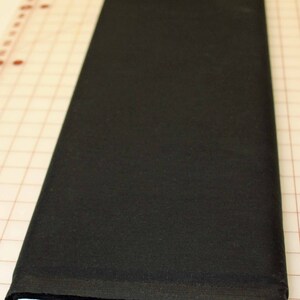 May include: A folded, solid black fabric roll. The fabric is tightly wound, revealing a crisp white edge. The fabric rests on a grid-patterned surface with inch measurements along the edge, suggesting it's for sewing or crafting.