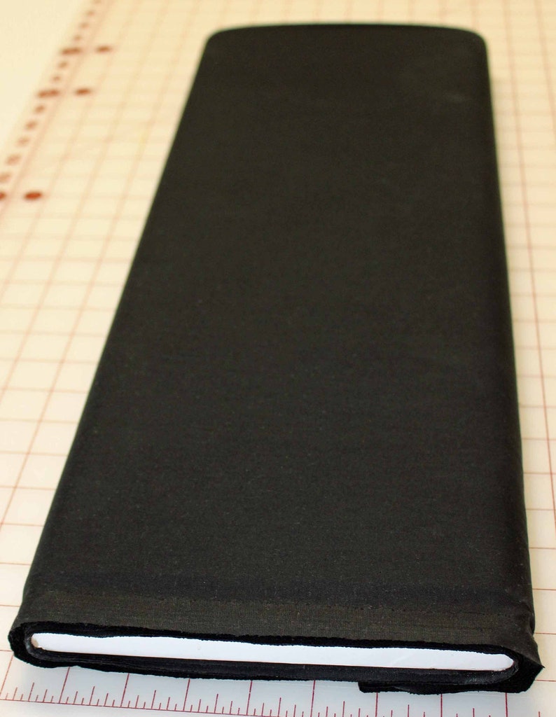 May include: A folded, solid black fabric roll. The fabric is tightly wound, revealing a crisp white edge. The fabric rests on a grid-patterned surface with inch measurements along the edge, suggesting it's for sewing or crafting.