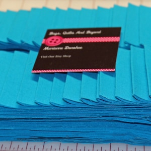 Puede incluir: Una pila de tiras de tela azul turquesa, dobladas y listas para coser. La tela est&aacute; dispuesta en un patr&oacute;n plisado. Una tarjeta de visita para "Big Quilts And Beyond" es visible en la parte superior de la tela.