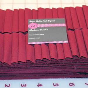 May include: A stack of burgundy fabric strips, folded and ready for sewing. The fabric is approximately 8 inches wide and is folded into 1/2 inch pleats. A business card for "Bags, Quilts and Beyond" is visible on top of the fabric.