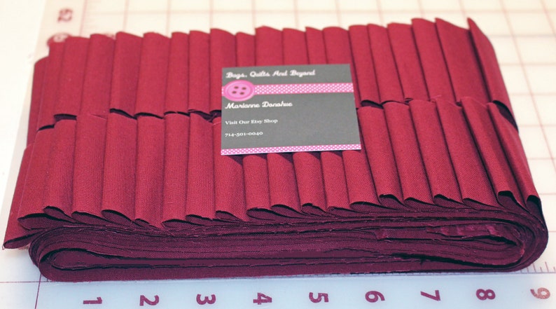May include: A stack of burgundy fabric strips, folded and ready for sewing. The fabric is approximately 8 inches wide and is folded into 1/2 inch pleats. A business card for "Bags, Quilts and Beyond" is visible on top of the fabric.