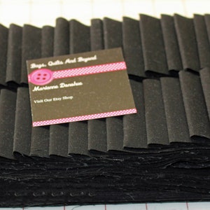 May include: A stack of black fabric strips, possibly for quilting or crafting. A business card with the text "Bags, Quilts And Beyond" and "Marianne Darahus" is visible. The fabric is folded in a layered pattern.