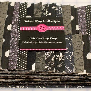 Puede incluir: Una pila de tiras de tela con varios patrones en negro, gris y blanco. Los patrones incluyen diseños florales, lunares y paisley. Una tarjeta de visita con el texto "Fabric Shop in Michigan" está encima.
