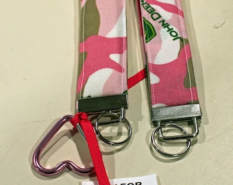 Puede incluir: Dos llaveros con un estampado de camuflaje rosa, blanco y verde. Cada llavero tiene un anillo de metal y una cinta roja. Un llavero tiene un cierre en forma de corazón. El texto "JOHN DEERE" es visible en los llaveros.