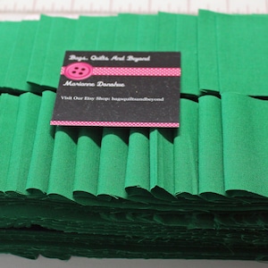 May include: A roll of green fabric with pleats, ready for sewing projects. The fabric is folded and stacked, with a business card on top that reads "Bags, Quilts And Beyond, Marianne Donahue, Visit Our Etsy Shop bagsquiltsandbeyond".