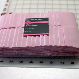 May include: A stack of folded pink fabric strips, possibly for quilting or sewing projects. A business card with a pink button logo and contact information rests on top of the fabric. The fabric is neatly arranged on a cutting mat.