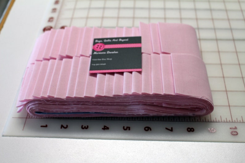 May include: A stack of folded pink fabric strips, possibly for quilting or sewing projects. A business card with a pink button logo and contact information rests on top of the fabric. The fabric is neatly arranged on a cutting mat.