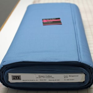 K&ouml;nnte beinhalten: Eine Rolle 100 % gek&auml;mmter Baumwolle in Wedgewood-Blau, 45 Zoll breit. Der Stoff ist mit "Dream Cotton" gekennzeichnet und stammt von Marshall Dry Goods Co., Inc. "Seit 1944" Batesville, AR 72501. Das Etikett enth&auml;lt auch eine Telefonnummer: 1-888-744-6277.