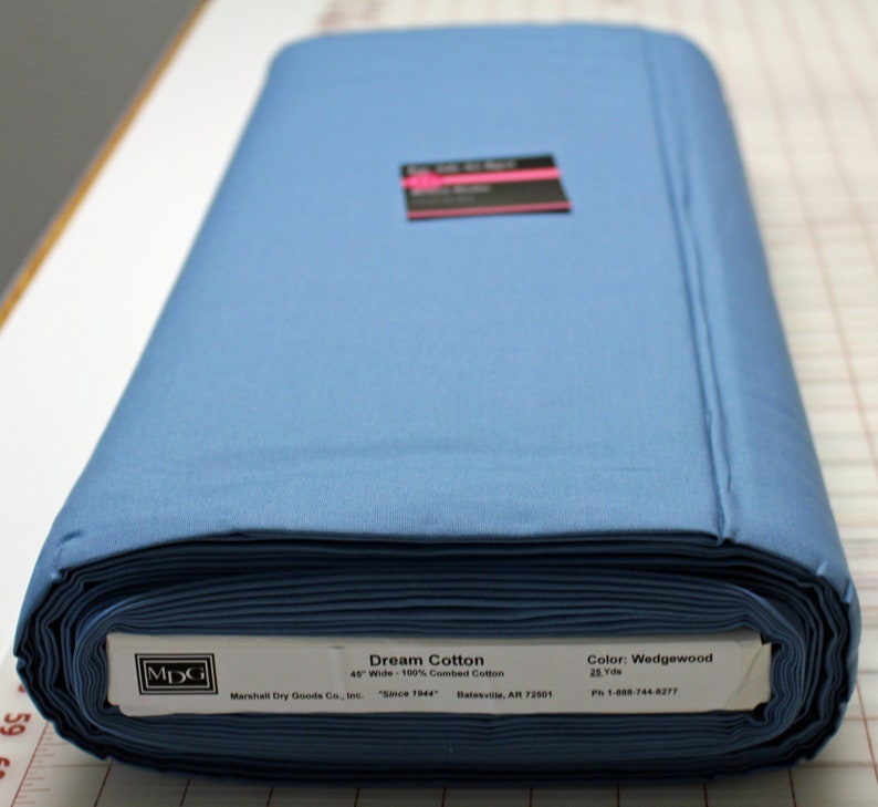 K&ouml;nnte beinhalten: Eine Rolle 100 % gek&auml;mmter Baumwolle in Wedgewood-Blau, 45 Zoll breit. Der Stoff ist mit "Dream Cotton" gekennzeichnet und stammt von Marshall Dry Goods Co., Inc. "Seit 1944" Batesville, AR 72501. Das Etikett enth&auml;lt auch eine Telefonnummer: 1-888-744-6277.