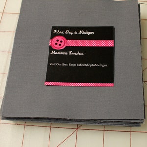May include: A stack of gray fabric squares with a black business card on top. The card features the text "Fabric Shop in Michigan," a pink button graphic, and the name "Marianne Danahue." The Etsy shop URL is also included.