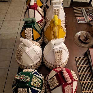 May include: A collection of handcrafted, decorative objects with a rounded base and a conical top. The objects feature a variety of colors, including white, yellow, green, and red, and are adorned with shells and beaded accents. The objects are arranged on a table.