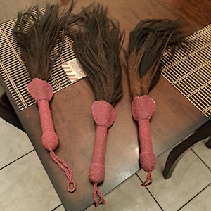 May include: Three long, dark brown hair whips with burgundy beaded handles. The whips are laid out on a wooden table with a woven mat.