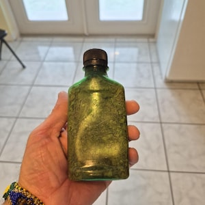 May include: A small, rectangular, translucent green glass bottle with a dark brown screw-top cap. The bottle is filled with a textured, dried green herbal substance that appears to be finely chopped or crushed leaves, filling the entire container.