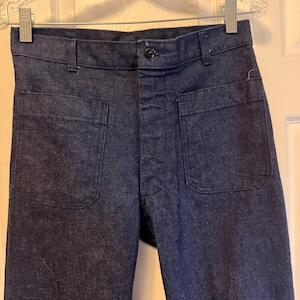 May include: Dark blue denim pants with two large front pockets and a button closure. The pants are hanging on a clear hanger. The denim has a slightly textured appearance.