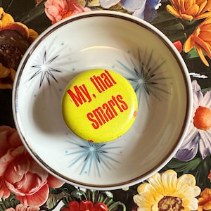 May include: A yellow button with the text "My, that Smarts" in red lettering rests inside a white bowl with a starburst design. The bowl is set against a floral background with various colorful flowers.