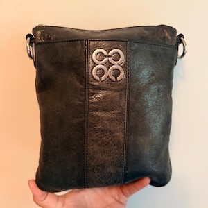 May include: A dark charcoal gray leather crossbody bag with a weathered texture. The front features a vertical center panel and a silver-toned metal logo consisting of four interlocking circles. It includes silver-toned hardware rings on the sides.
