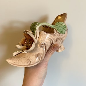 May include: A ceramic shoe-shaped vase with a floral design. The vase is painted in shades of pink and brown with green leaves and a white flower.